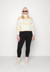 Sweatshirt em creme com tie-dye laranja, punhos canelados, combinado com jeans skinny pretos e botas de combate bege. Modelo está de pé contra um fundo branco.