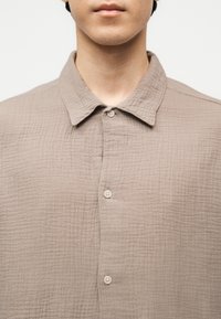 Κοντινό πλάνο ατόμου που φοράει μπεζ υφασμάτινο πουκάμισο με κουμπιά και γιακά, εμφανίζοντας το κάτω μέρος του προσώπου και το λαιμό.