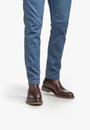 Botas Chelsea de cuero marrón con un acabado texturizado, combinadas con pantalones ajustados de color azul claro, que presentan un dobladillo simple. La suela tiene un diseño con relieve.