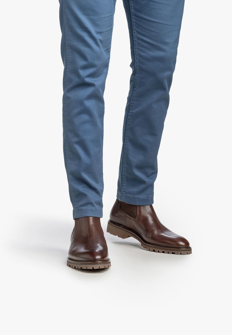 Botas Chelsea de cuero marrón con un acabado texturizado, combinadas con pantalones ajustados de color azul claro, que presentan un dobladillo simple. La suela tiene un diseño con relieve.