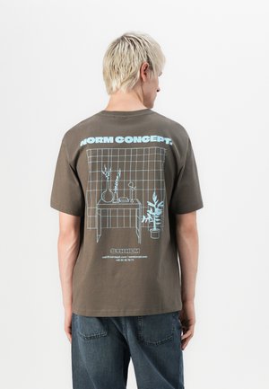 Brun bomull t-shirt med en grafisk design i ljusblått, som skildrar ett bord med en vas och växt mot ett rutmönster på baksidan.