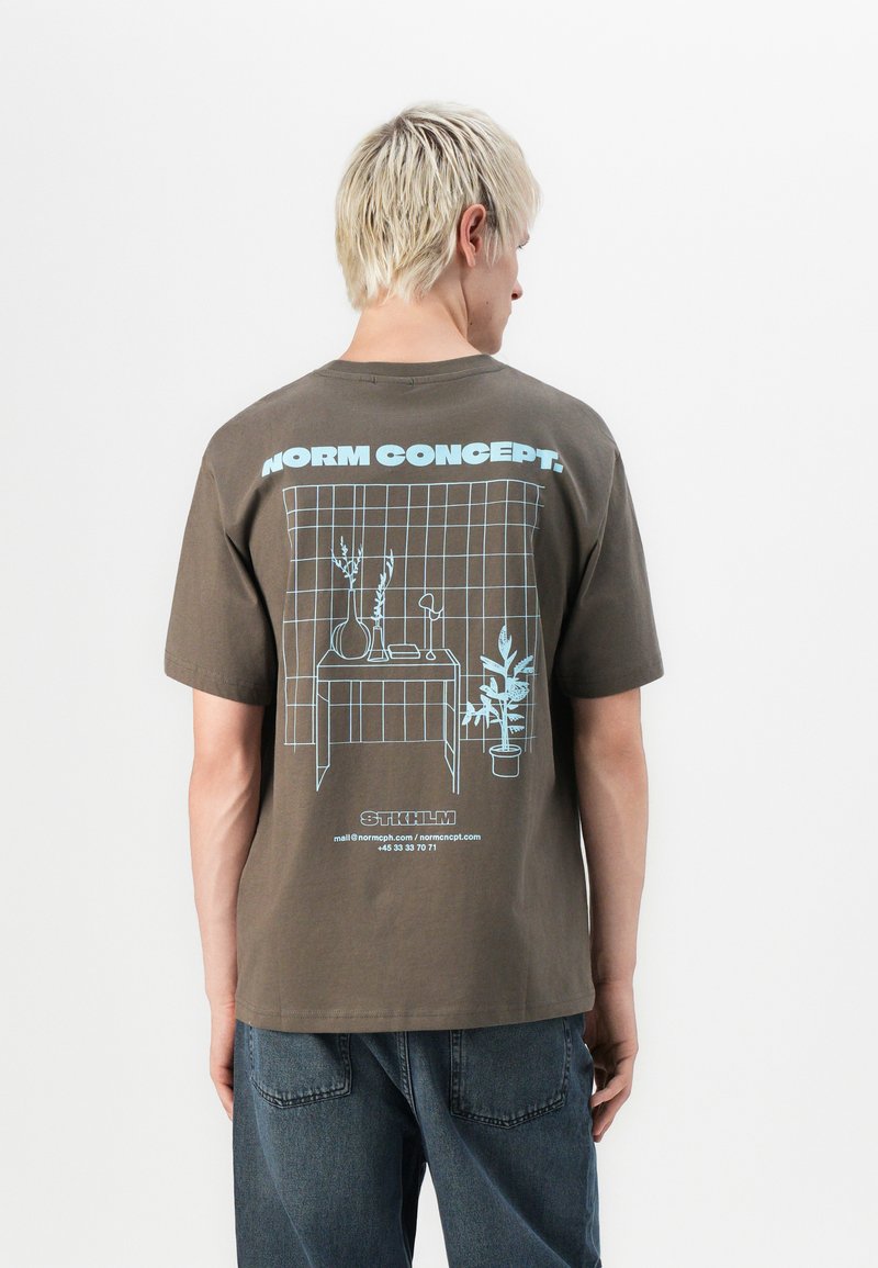 Bruine katoenen t-shirt met een grafisch ontwerp in lichtblauw, waarop een tafel met een vaas en plant staat afgebeeld, tegen een gridpatroon op de achterkant.
