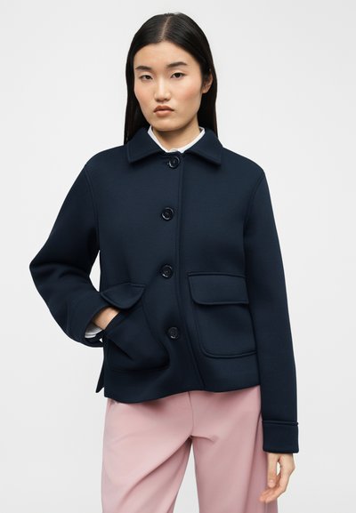 Γυναίκα που φοράει μπλε-ναυτικό μπουφάν με μεγάλες μπροστινές τσέπες και ανοικτό ροζ παντελόνι, στέκεται μπροστά σε απλό φωτεινό φόντο.