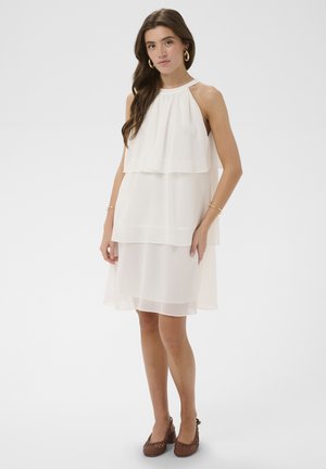 Naine, kes kannab valget varrukateta kihilist kleiti, kuldseid rõngaskõrvarõngaid, käevõrusid ja pruune punutud kõrgkontsaga kingi, seisab valge tausta ees.