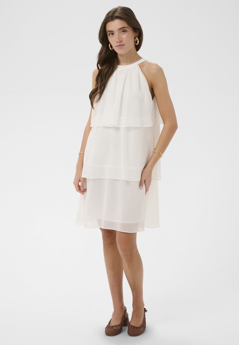 Frau trägt ein ärmelloses weißes, geschichtetes Kleid mit goldenen Creolen, Armbändern und braunen, geflochtenen High Heels, steht vor weißem Hintergrund.