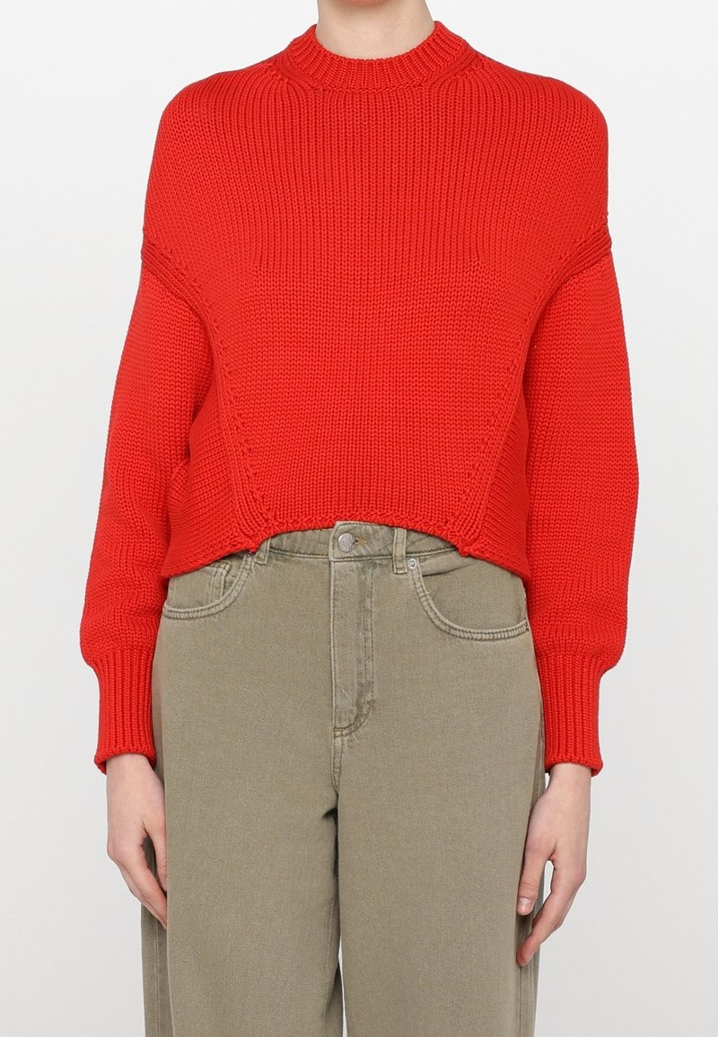 Personne portant un pull tricoté rouge vif à manches longues et un pantalon marron clair taille haute, debout devant un fond uni.