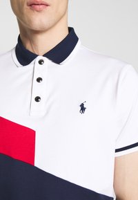 Polokošile se bílým tělem, námořnickým límcem, červeno-námořnickým horizontálním barevným blokem a černými knoflíky s vyšitým logem.