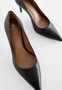 Zapatos de tacón alto negros de punta afilada, hechos de cuero liso, con un diseño cónico y un tacón fino tipo stiletto. Textura del plantillón en un tono marrón claro es visible.