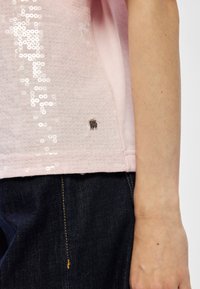 Gros plan d'une personne portant un haut rose clair à sequins et un jean bleu foncé, se concentrant sur l'ourlet latéral inférieur et le bras.
