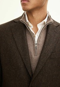 Blazer a spina di pesce marrone su un maglione grigio con zip, abbinato a una camicia bianca con colletto. I materiali strutturati creano un look stratificato.