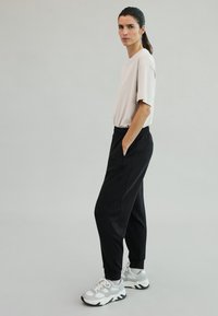 Light beige t-shirt with short sleeves, paired with black elastic waistband pants. Model wears gray and white sneakers, standing on a plain background.