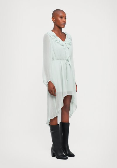 Rochie de chiffon verde deschis cu guler volanat și talie cu cravată, având un hem asimetric. Asortată cu cizme negre cu toc până la genunchi.
