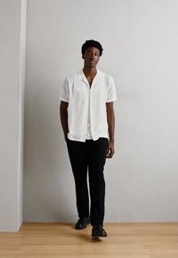 Cămașă albă cu mânecă scurtă și nasturi, cu un model texturat, asociată cu pantaloni negri și încălțări închise, pe o persoană care stă lângă un zid deschis la culoare.