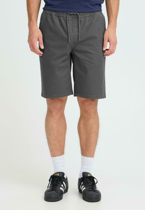 Grau Stoff-Shorts mit elastischem Bund und Kordelzug, die bis zur Mitte des Oberschenkels reichen. Kombiniert mit schwarzen Turnschuhen und weißen Socken.