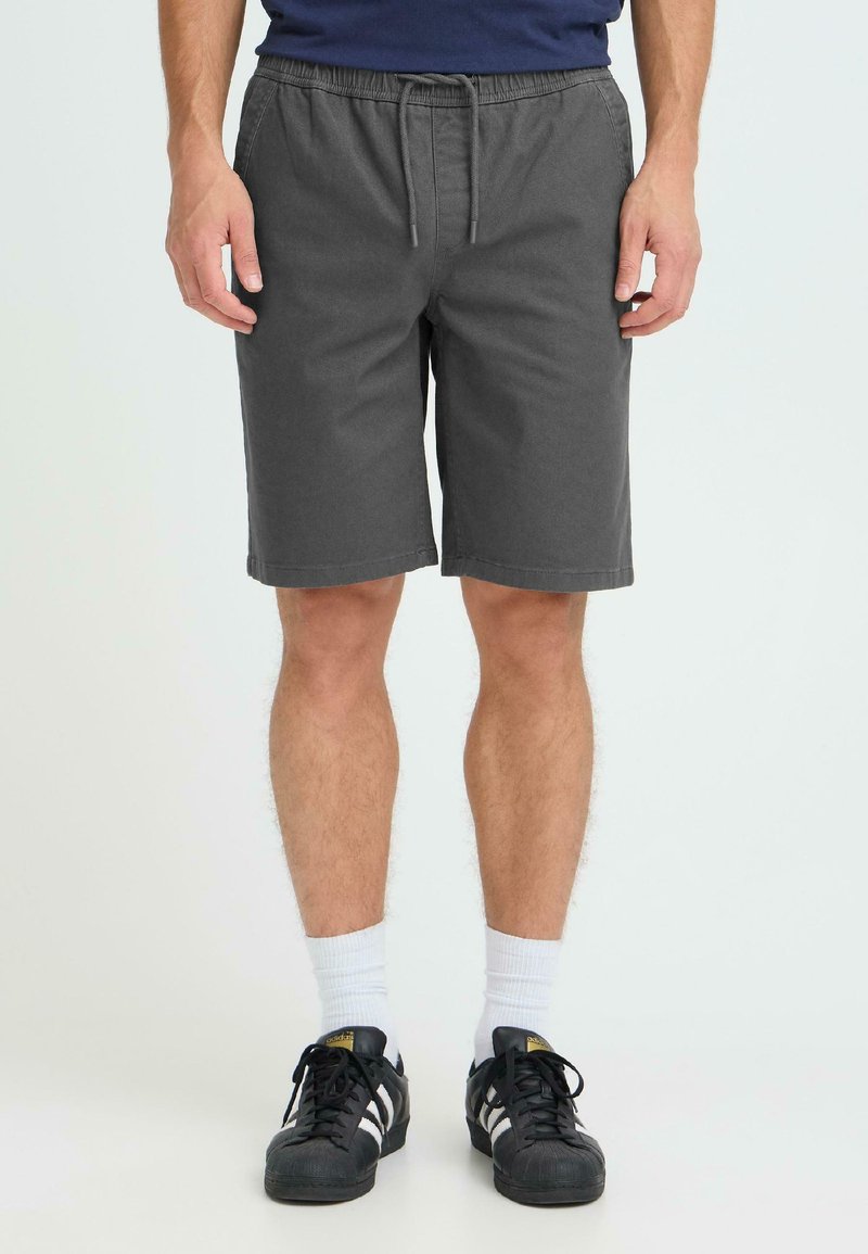 Shorts gris en tissu, dotés d'une ceinture élastique avec cordon de serrage, arrivant à mi-cuisse. Portés avec des baskets noires et des chaussettes blanches.