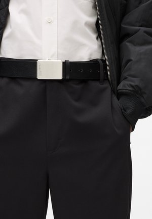 Černý kožený pásek se stříbrnou obdélníkovou kovovou sponou, nošený s bílou košilí a černými kalhotami, které mají jemné detaily kapes.