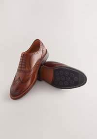 Chaussures habillées en cuir marron avec perforations décoratives, bout rond et laçage fermé. Semelle en caoutchouc noire avec motif de bande de roulement circulaire.