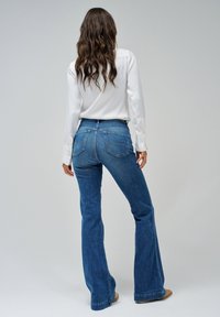 Jeans in denim svasato di blu medio, con tasche posteriori con dettagli di cucitura, abbinati a una camicetta bianca a maniche lunghe.