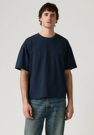Ung mand iført en simpel mørk navy oversized t-shirt og blå jeans, stående mod en lysegrå baggrund.
