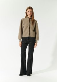 Beige gebreide cardigan met een hoge kraag, gegolfde randen en knopen aan de voorkant, gecombineerd met zwarte uitlopende jeans met zichtbare stiksels.