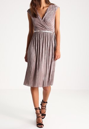 Kobieta w plisowanej, połyskującej mauve sukience kopertowej z cienkim białym paskiem i czarnych sandałach na obcasie z wiązaniami.