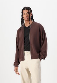 Brun zip-up bomberjakke med en avslappet passform, brukt over en svart t-skjorte. Kombinert med lys beige, lette bukser.