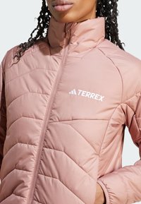 Veste Terrex rose clair avec un col montant, un motif matelassé, une fermeture éclair et des détails de logo blancs sur la poitrine et l'épaule.