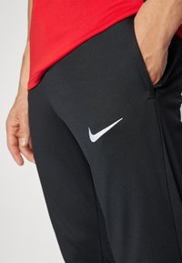 Schwarze Sporthose mit glatter Textur, ausgestattet mit einem weißen Nike Swoosh-Logo. Kombiniert mit einem roten Shirt, die Hand liegt in der Tasche.