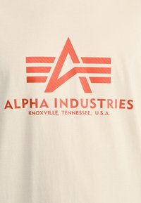Cream t-shirt featuring a red graphic design with an angular logo and stripes. Text reads "ALPHA INDUSTRIES, KNOXVILLE, TENNESSEE, U.S.A."