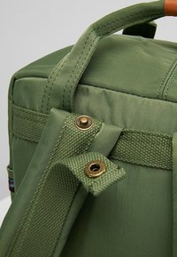 Zelený textilní batoh se strukturovaným tvarem, s texturovanými popruhy, bronzovými kovovými prvky a švy.