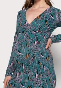 Turkos klänning med långa ärmar och djup V-ringning. Har ett abstrakt mönster i rosa, brunt och svart. Elastiskt tyg för komfort.