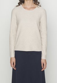 Pull en maille côtelée beige à manches longues avec un col rond, porté avec une jupe bleu marine foncé, offrant une coupe décontractée et un tissu texturé.