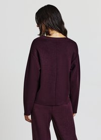 Bordeauxroter Strickpullover mit lockerem Schnitt, Bootsausschnitt und überschnittenen Schultern. Nahtdetail auf der Rückseite, weiche Textur und lange Ärmel.