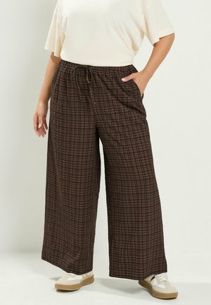 Person trägt braune karierte Hose mit weitem Bein und Kordelzug in der Taille, weißes kurzärmeliges Oberteil und weiße Sneakers, steht mit einer Hand in der Tasche.