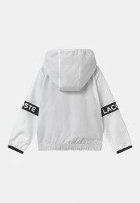 White hooded jacket with black accents. Features elastic cuffs and a drawstring hem. "LACOSTE" logo displayed prominently on sleeves.