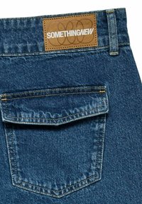 Vaqueros de denim con un lavado azul oscuro, que presentan una etiqueta de cuero marrón cosida en la cintura y un solo bolsillo trasero con costuras de contraste.