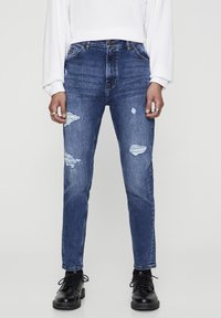 Vaqueros de mezclilla azul con un ajuste slim, que presentan desgastes y rotos en los muslos y las rodillas. Combinados con una camisa blanca de manga larga y botas negras.