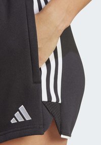 Schwarze Sportshorts mit weißen Seitenstreifen, ausgestattet mit einer Tasche und einem silbernen Adidas-Logo. Der Stoff wirkt glatt und leicht glänzend.