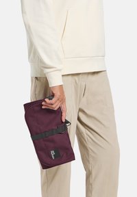 Sacchetto di tessuto bordeaux con forma curvata, cinturino e hardware neri. Marchio visibile. Tenuto a mano, indossato con abbigliamento di colori chiari.