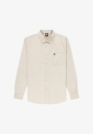 Camisa de botones de manga larga color beige, hecha de tela de pana, con un bolsillo en el pecho y una etiqueta de marca en el cuello.