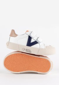 Sneakers bianchi per bambini con dettagli blu scuro, dotati di cinturini in velcro, una suola in gomma texturizzata e una linguetta sul tallone beige. Logo stampato sul lato.
