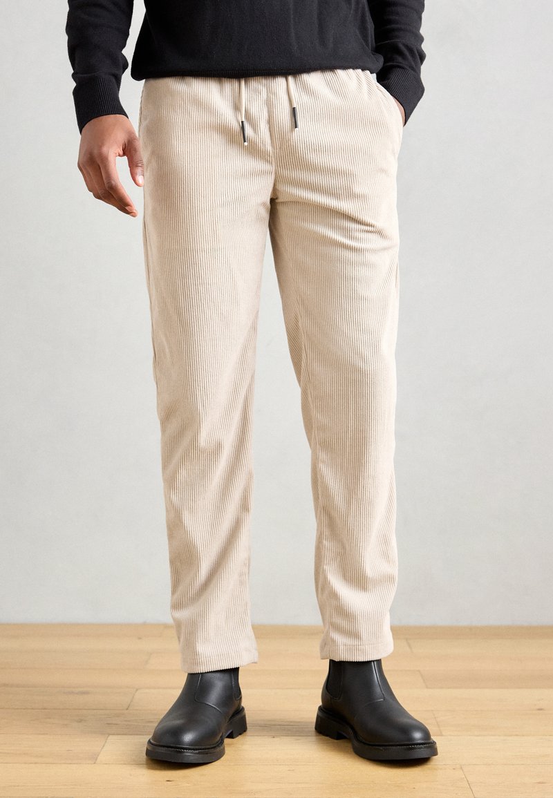 Pantalon en velours côtelé beige à coupe décontractée, avec une taille à cordon et des rayures verticales subtiles, associé à des bottines noires.