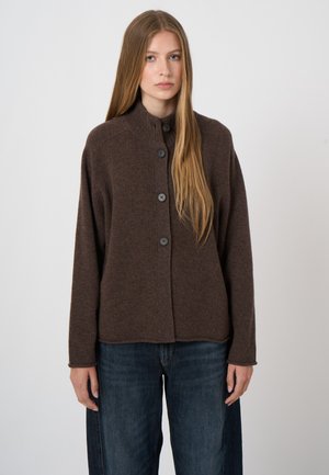 Jonge vrouw met lang blond haar die een bruine vest met knopen en donkerblauwe spijkerbroek draagt, staand tegen een effen lichte achtergrond.