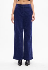 Πανάκριβα, ψηλόμεσα παντελόνια από χοντρό βελούδο σε ναυτικό μπλε, συνδυασμένα με μαύρες γωνιακές μπότες μέχρι τον αστράγαλο, προβαλλόμενα από τη μέση μέχρι τα πόδια.