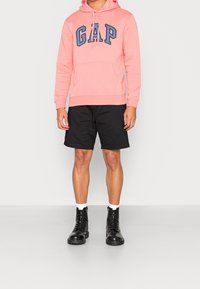 Persona vestida con una sudadera rosa de GAP, pantalones cortos negros, calcetines blancos y botas negras de cordones, de pie frente a un fondo sencillo.
