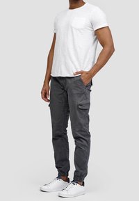 Weißes T-Shirt mit Rundhalsausschnitt und Tasche, kombiniert mit grauen Cargo-Hosen mit Seitentaschen und schmal zulaufenden Säumen. Getragen mit weißen Sneakern.
