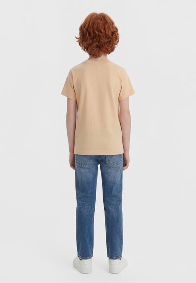 Niño con cabello rizado y rojo de espaldas, vistiendo una camiseta de manga corta beige, jeans azules y zapatillas blancas sobre un fondo neutro.