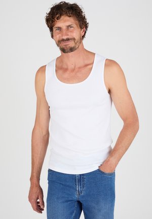 Hombre con cabello rizado y barba, vestido con una camiseta sin mangas blanca y jeans azules, de pie con una mano en el bolsillo contra un fondo liso.