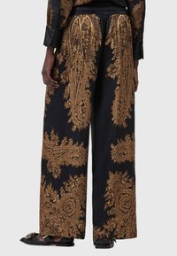 Laiad püksid mustast kangast, millel on keerukad kuldsed paisleimustrid, elastne vöökoht ja lõtv istuvus. Täiendavad lamedad mustad sandaalid.