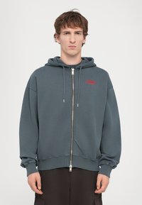 Haut à capuche gris à fermeture éclair avec une coupe décontractée, grande capuche et texte rouge "Snake Charmer". Présente des poignets côtelés et une texture lisse.